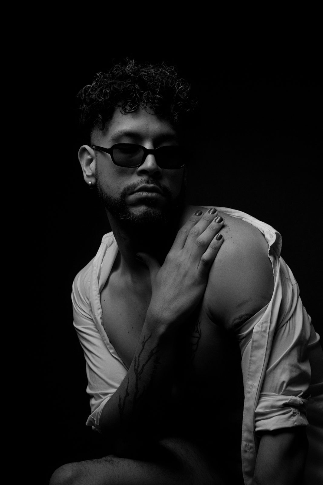 Retrato en blanco y negro de un hombre con gafas de sol y camisa abierta, con la mano sobre el hombro.