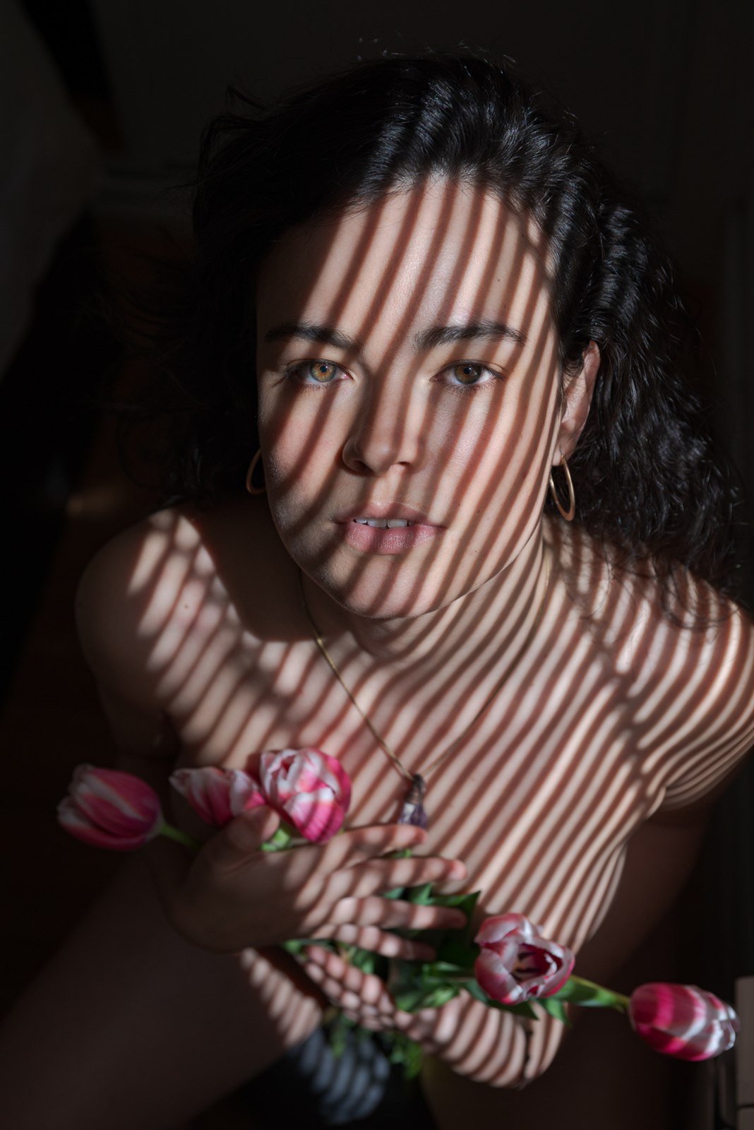 Retrato en color con franjas de luz de persiana sobre el rostro y los hombros, con un ramo de tulipanes en las manos.