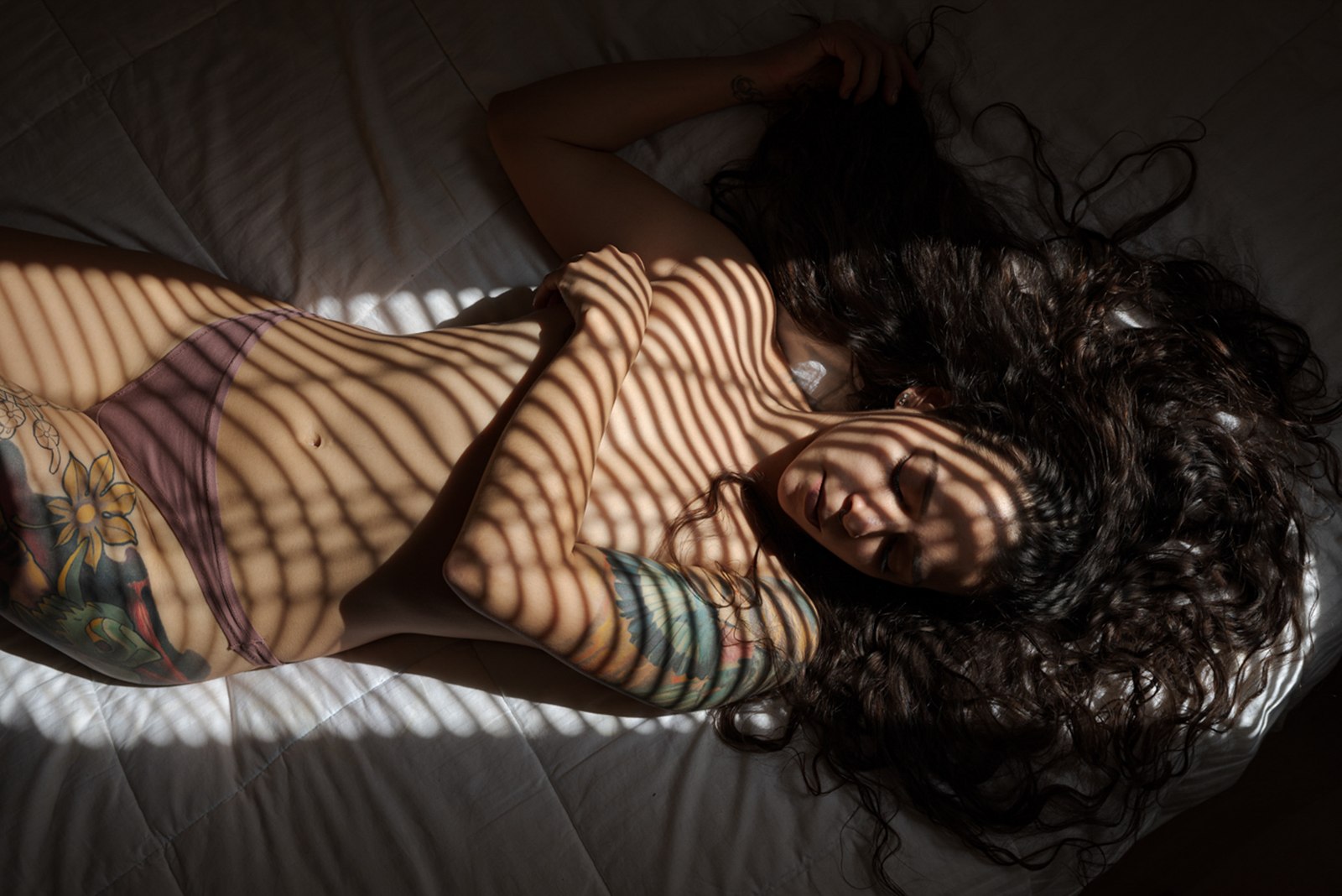 Vista cenital de una mujer tumbada en una cama, con los ojos cerrados y el pelo suelto, con franjas de luz de persiana sobre el cuerpo.