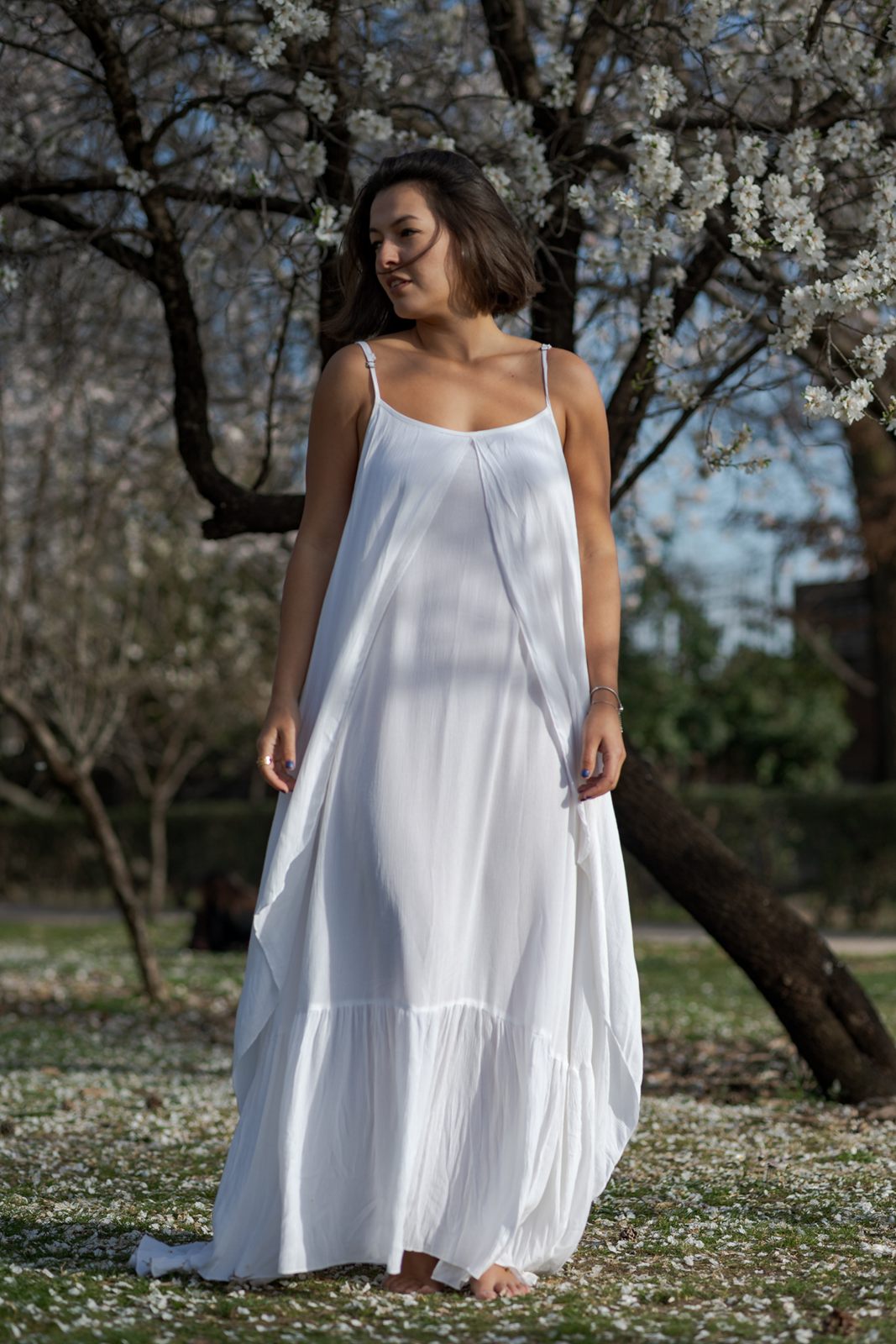 Mujer con vestido blanco bajo un almendro en flor, mirando hacia un lado.
